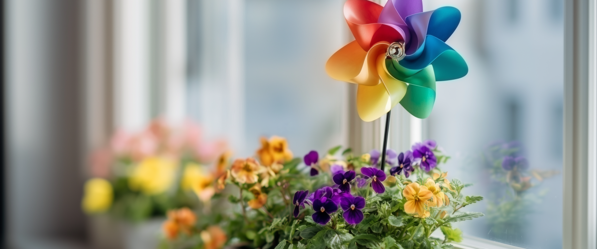 Bunter Fensterkasten mit Stiefmütterchen und Windrad in Regenbogenfarben, auf einer Fensterbank eines modernen Hauses als Teil eines umweltfreundlichen Hausbaus mit Fokus auf urbane Begrünung.
